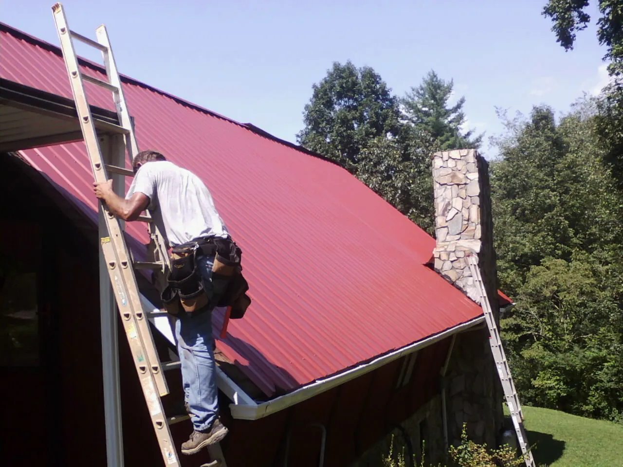 Roofer on a ladder beside a red metal roof and stone chimney — Soffit & Fascia Repair by Roofing Brothers in Mukilteo